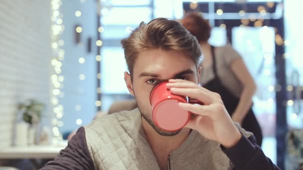 Молодой человек смотрит в камеру и пьет кофе в кафе — стоковое видео