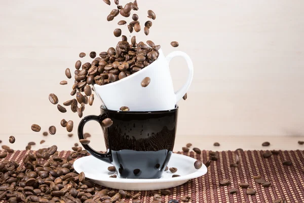 Granos de café tostados cayendo en una taza blanca de pie sobre una taza negra sobre un plato blanco de pie sobre una manta sobre un fondo de madera claro . —  Fotos de Stock