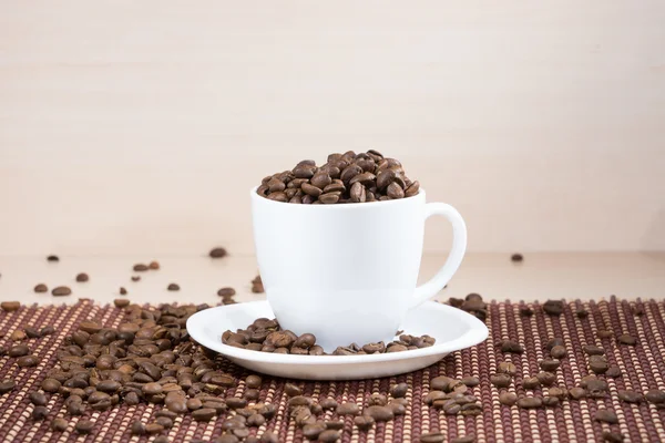 Taza blanca llena de granos de café tostados de pie sobre un plato blanco sobre mantel sobre fondo de madera clara . —  Fotos de Stock