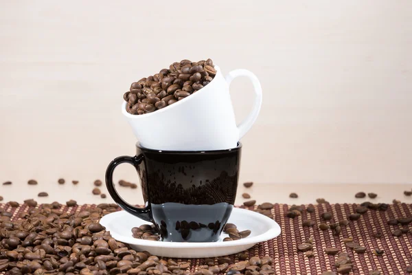 Copa blanca llena de granos de café tostados de pie en una taza negra en un plato blanco de pie sobre una manta sobre fondo de madera clara . —  Fotos de Stock