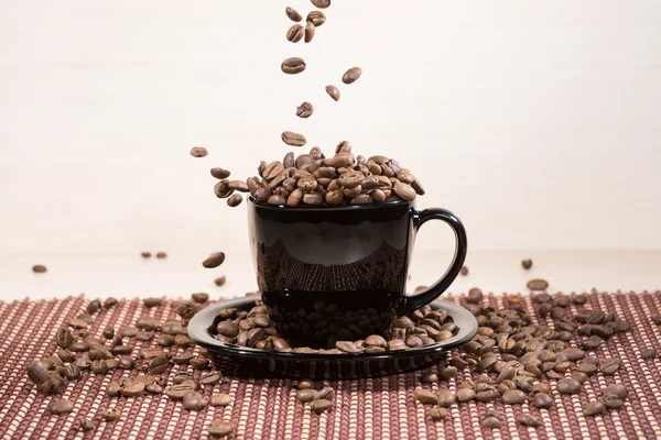 Vista de la taza negra de pie en el plato negro con la caída de granos de café tostados en la tabla . —  Fotos de Stock
