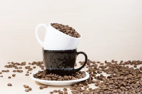 Copa blanca de pie en una taza negra en plato blanco de pie sobre fondo de madera clara con lleno de granos de café tostados en la mesa . —  Fotos de Stock