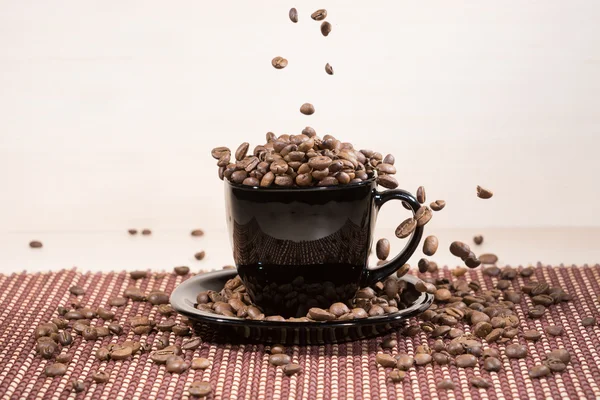 Granos de café tostados cayendo en una taza negra de pie sobre un plato negro de pie sobre una manta . —  Fotos de Stock