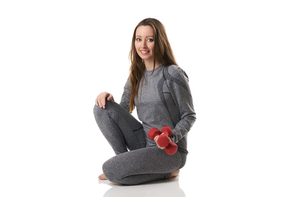Beautiful woman in sitting position keeping two red dumbbells in one hand in gray thermal underwear. — Stock Photo, Image