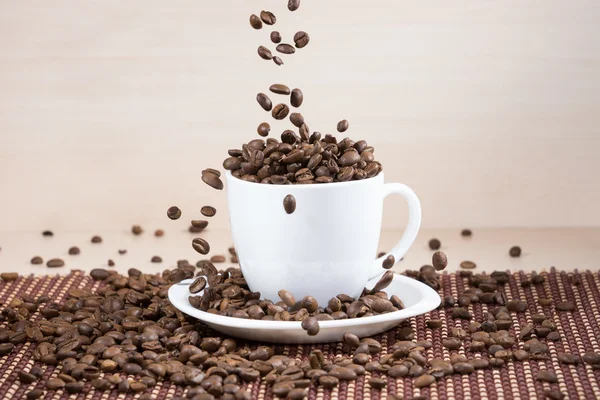 White cup full of brown roasted coffee beans on plate with falling beans from top on tablemat.