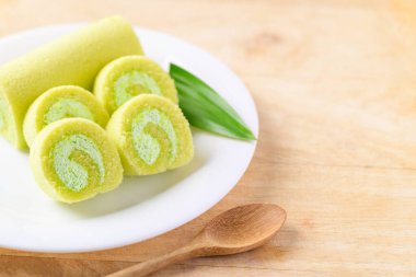 Dilimlenmiş Pandan pastası, beyaz tabakta pandan yaprağı ve tahta masada kaşık.