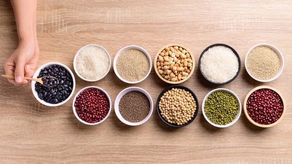 Various cereal grain in a bowl with hand on wooden background (quinoa seeds, black kidney bean, peanut, soybean, azuki beans, rice grain and mung beans)