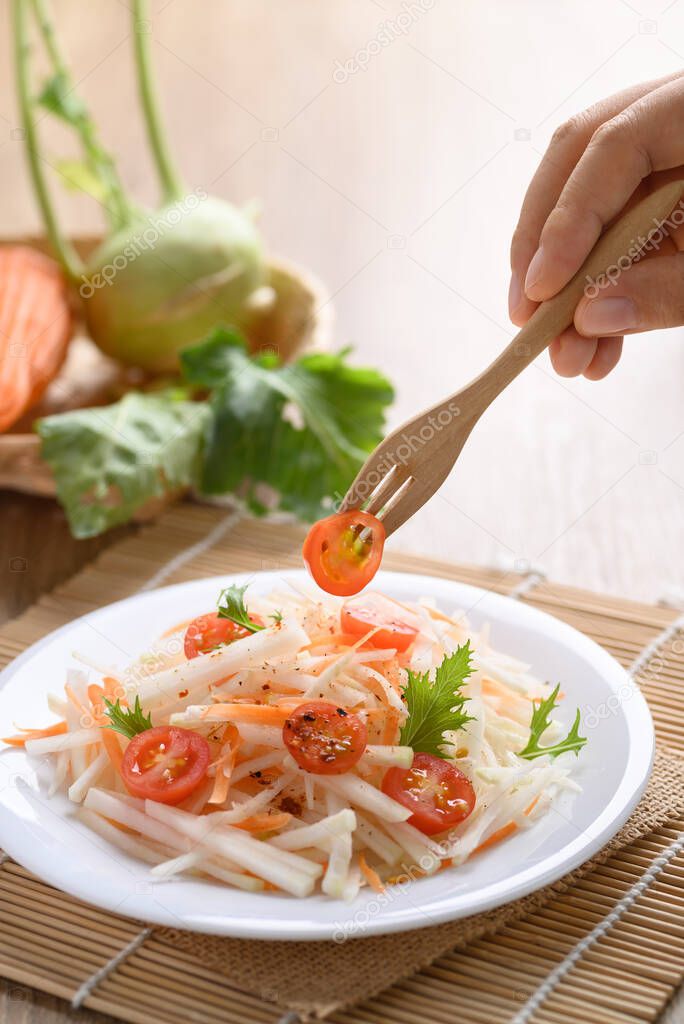 Hand holding fork and eating salad with sliced kohlrabi, carrot, tomato and mizuna leaf on white dish, Vegan food, Healthy food