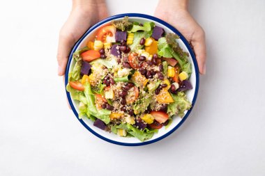 Marul, kinoa tohumu, azuki fasulyesi, mısır, domates, balkabağı ve mor patatesli sağlıklı sebze salatası beyaz arka planda elle tutulan bir kasede. Vejetaryen ve bitkisel gıda