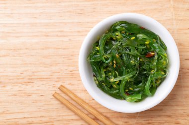 Tahta zemin üzerinde çubuklar olan bir kasede susam tohumlu yosun salatası, masa üstü görünümü.