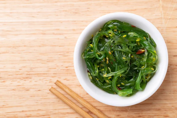 Tahta zemin üzerinde çubuklar olan bir kasede susam tohumlu yosun salatası, masa üstü görünümü.