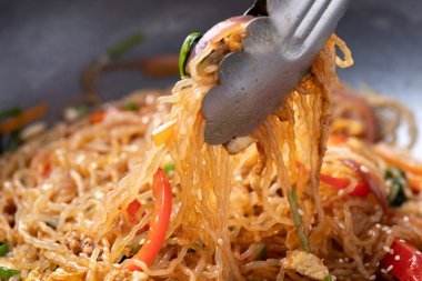 Japchae, Korean stir fried glass noodles with vegetables, pork, soy sauce and sesame oil in cooking pan and food tongs