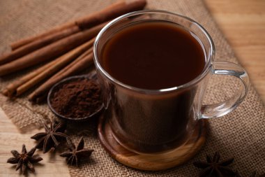 Hot cocoa drink in cup glass with cocoa powder, cinnamon and star anise on brown background