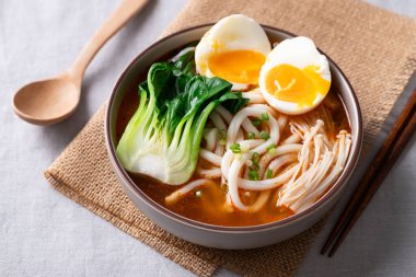 Baharatlı Udon Erişte Çorbası 'nın üstünde Bok Choy, Enoki Mantarı ve yemek çubukları ve kaşık kaplı yumuşak kaynamış yumurta var.