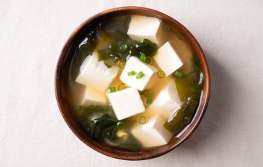 Sağlıklı Japon miso çorbasının üst görüntüsü yumuşak tofu ve yosun ile ahşap bir kasede beyaz kumaş arka planda.