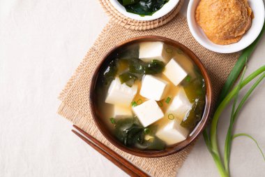Sağlıklı Japon miso çorbasının üst görüntüsü yumuşak tofu ve yosun ile ahşap bir kasede beyaz kumaş arka planda yemek çubukları ile