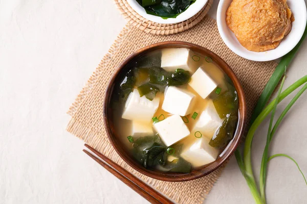 Sağlıklı Japon miso çorbasının üst görüntüsü yumuşak tofu ve yosun ile ahşap bir kasede beyaz kumaş arka planda yemek çubukları ile