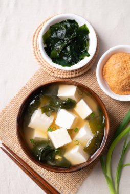 Sağlıklı Japon miso çorbasının üst görüntüsü yumuşak tofu ve yosun ile ahşap bir kasede beyaz kumaş arka planda yemek çubukları ile