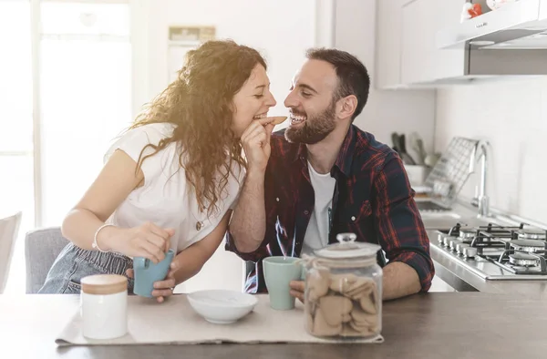 Engaged couple has breakfast together in their new home - young couple jokes while eating cookies - love and well-being concept - warm filter on background