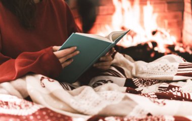Soğuk bir günde kadın dinleniyor ve ısınıyor. Battaniyeyle örtüyor ve kitap okuyor. Arkasında şömine olan neşeli bir atmosfer.