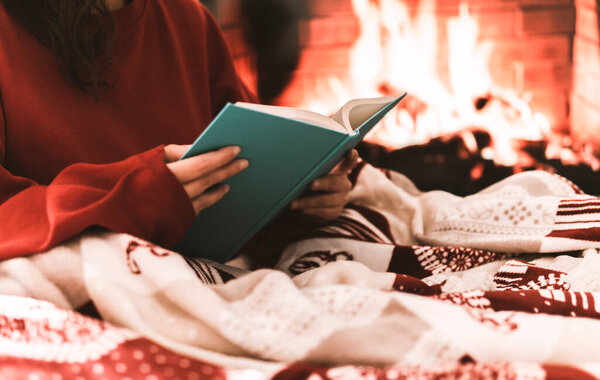 Female resting and warming up in a cold day covering with a blanket and reading a book - festive atmosphere with fire place on background