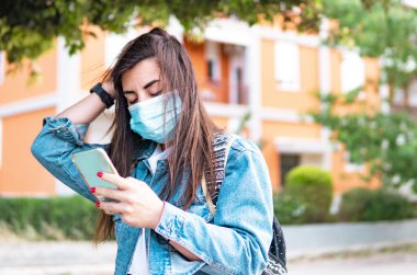 Koruyucu maskeli genç bir kız sırtında sırt çantasıyla gezen bir kadın koronavirüs salgını sırasında akıllı telefonun yönünü kontrol ediyor.