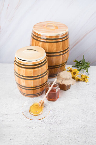 Two ceramic barrels and two glass jars with honey and a saucer with honey and a wooden spoon on a light background. Copy space