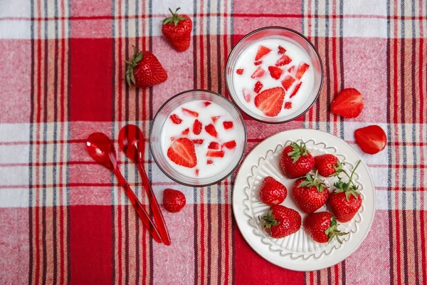 Two Glasses of Yoghurt,Red Fresh Strawberries are in the Ceramic Plate with Plastic Spoons on the Check Tablecloth.Breakfast Organic Healthy Tasty Food.Cooking Vitamins Ingredients.Summer Fruits. — Stock Photo, Image