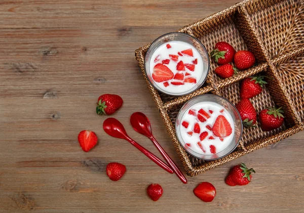 Zwei Gläser Joghurt, rote frische Erdbeeren in der Rattanbox mit Plastiklöffeln auf dem Holztisch. Frühstück bio, gesund und schmackhaft mit Vitaminen. Sommerfrüchte. — Stockfoto