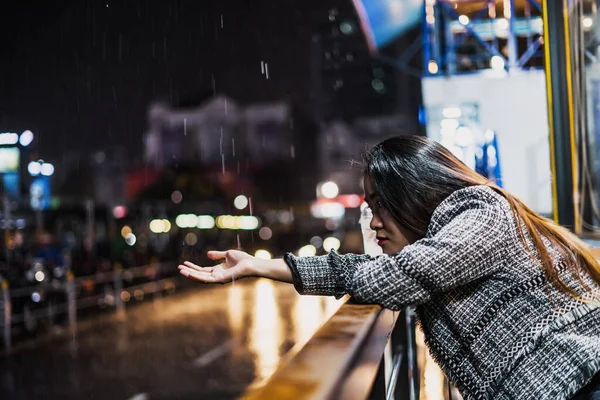 beautiful young lonely asian girl standing outside a cafe or restaurant, on a street night, it's raining, she is waiting for a bus or someone meets. Selective focus.