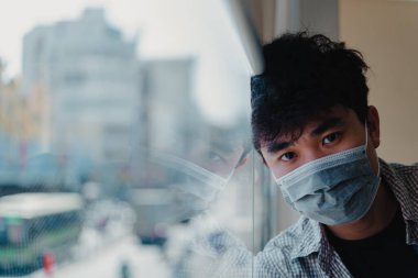 Yüz maskesi takan Asyalı genç adam COVID19 salgını nedeniyle kendini karantinaya almak için evde karantinada kalıyor. Pencereden dışarı bakıyor. Üzgün yüz ve gözler. Seçici odak.