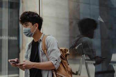 Açık havada akıllı telefon kullanarak cerrahi maske takan Asyalı bir adam. Sağlık ve tıbbi konsept