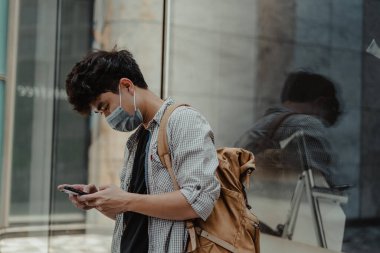 Açık havada akıllı telefon kullanarak cerrahi maske takan Asyalı bir adam. Sağlık ve tıbbi konsept