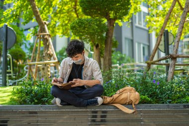 Koruyucu maskeli genç Asyalı bir adam salgın sırasında dışarıda deri deftere yazı yazıyor. Sağlık ve Eğitim Konsepti.