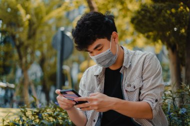 Maske takan ve akıllı telefon kullanan, arama yapan ya da sosyal medya konsepti olan genç Asyalı bir adam arkadaşlarına mesaj yazıyor. Seçici odak.