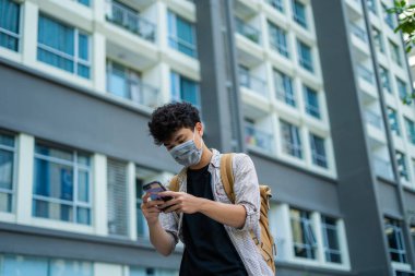 Young asian man wearing mask and using smartphone, searching or social networks concept, man typing an sms message to his friends in outdoor. Selective focus. clipart