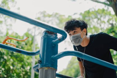 Sosyal uzaklık. Korumalı maskeli genç Asyalı adam dışarıda parkta çalışıyor. Şınav çekerek ve barfiks çekerek. Karantinada spor. Covid-19. Korunma. Seçici odak.