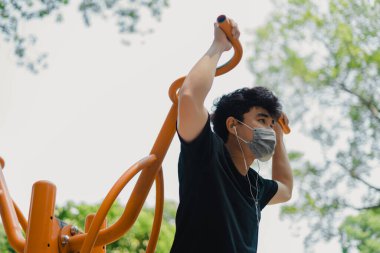 Sosyal uzaklık. Korumalı maskeli genç Asyalı adam dışarıda parkta çalışıyor. Şınav çekerek ve barfiks çekerek. Karantinada spor. Covid-19. Korunma. Seçici odak.