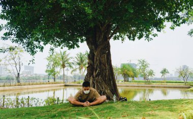 Yüzünde koruyucu maskeyle meditasyon yapan Asyalı bir adam covid-19 'un kilit altında büyük bir ağacın altında yoga egzersizi yapıyor. Seçici odaklanma. Sosyal uzaklık. Karantinada spor.