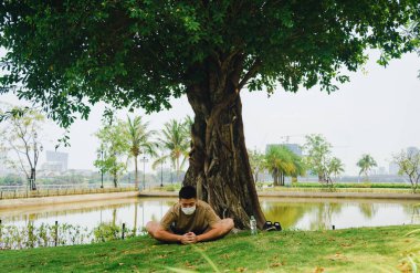 Yüzünde koruyucu maskeyle meditasyon yapan Asyalı bir adam covid-19 'un kilit altında büyük bir ağacın altında yoga egzersizi yapıyor. Seçici odaklanma. Sosyal uzaklık. Karantinada spor.