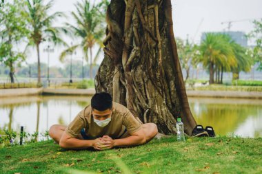 Yüzünde koruyucu maskeyle meditasyon yapan Asyalı bir adam covid-19 'un kilit altında büyük bir ağacın altında yoga egzersizi yapıyor. Seçici odaklanma. Sosyal uzaklık. Karantinada spor.