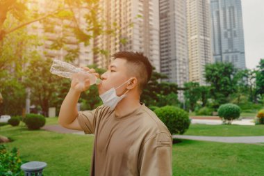 Koşmayı ve çalışmayı bitirdikten sonra su içerken yüz maskesi takan Asyalı genç koşucu. Virüsü ve alerji enfeksiyonunu korumak için maske takıyor..