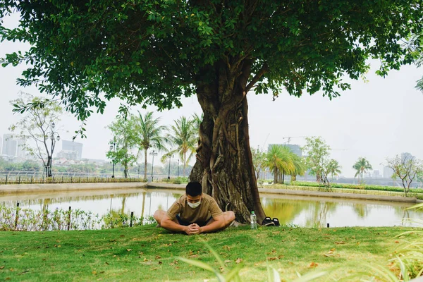 Asian man practicing meditation with wear protective mask on face, yoga exercise under big tree in park during lock down of covid-19. Selective focus. Social distancing. Sport in quarantine.