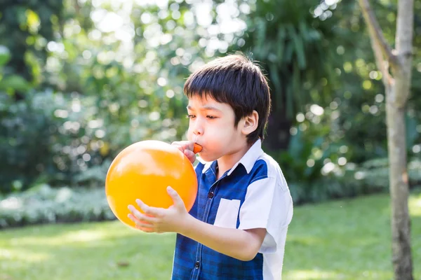 Niño soplando un globo naranja en el parque en un día soleado . — Foto de Stock