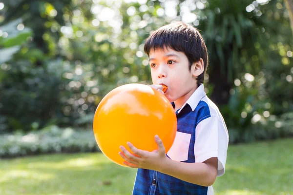 Niño soplando un globo naranja en el parque en un día soleado . — Foto de Stock