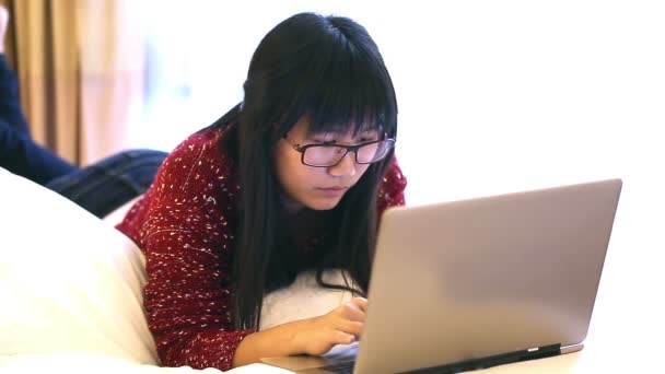 Asiática mujer usando gafas acostada en una cama en casa y ella trabaja en un ordenador portátil — Vídeos de Stock