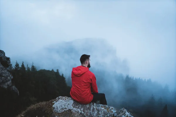 年轻的留着胡子的徒步旅行者穿着红色夹克 望着山顶和森林 周围有雾和雾气 在罗马尼亚Ceahlau Massif 徒步旅行者站在山顶欣赏风景的背景 — 图库照片