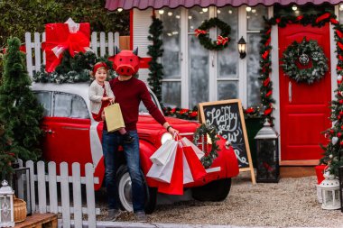 Kırmızı boğa maskeli genç bir adam küçük bir bebek tutuyor, beyaz paketler. Yeni yıl alışverişi, indirim. Aile arabanın yanında duruyor, Noel için kardaki ağaçlar süslenmiş. Kız öküzü öpüyor. Boşluğu kopyala
