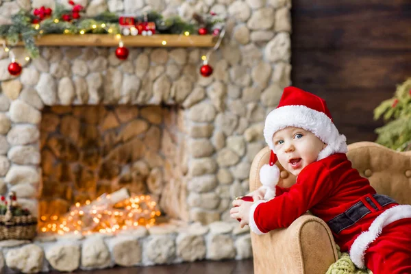 赤いサンタクロースの服を着た小さな男の子のクローズアップ肖像画 明るいガーランドと石の暖炉で遊んで クリスマスツリーの下にギフトボックスを置きます 年賀状 赤ん坊は笑い 楽しみを持っている — ストック写真