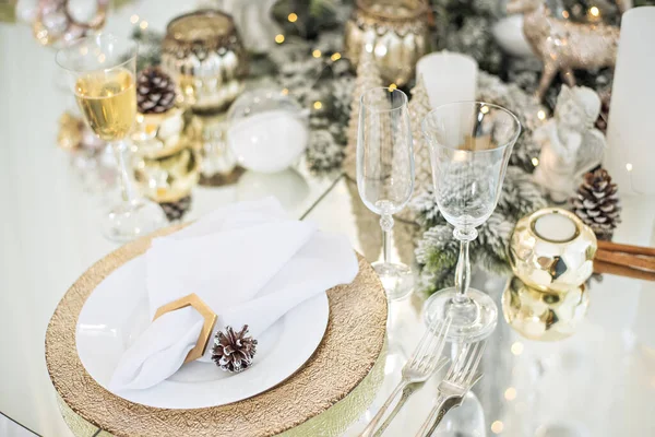 New Year\'s festive glass table setting. Greeting card. In the branches of the Christmas tree are champagne glasses, cutlery, a plate with gilding, a napkin ring. Decorated with gold candle. Copy space